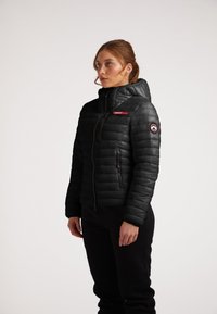 Giubbotto imbottito nero con zip frontale, cappuccio e un'etichetta rosso accentuata. Presenta un logo rotondo sulla manica e una superficie liscia.