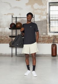 Camiseta preta de algodão, calções bege claros, meias pretas e ténis brancos. A carregar um saco de desporto preto; parede de tijolos ao fundo com prateleiras.