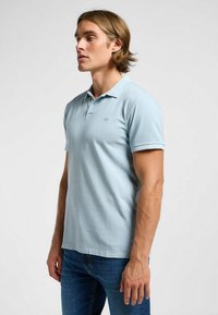 Ljusblå polo t-shirt i bomull, med kort krage, korta ärmar och en liten logotyp på vänster bröst. Bärs med blå jeans.