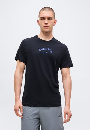 Νέος άνδρας με μαύρο T-shirt "Chelsea" της Nike και γκρι σορτς Nike Pro, στέκεται μπροστά σε απλό ανοιχτό φόντο.