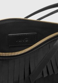 Interno di una borsa nera con zip dorata, caratterizzata da un tessuto testurizzato e un'etichetta in pelle con inciso 'MNG'. Presenta dettagli di design con frange.