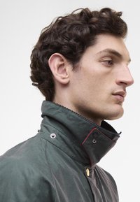 Σκούρο πράσινο μπουφάν με ψηλό γιακά, διακοσμημένο με ασημένια κουμπιά και κόκκινες ραφές, κατασκευασμένο από υφασμάτινο υλικό με υφή.