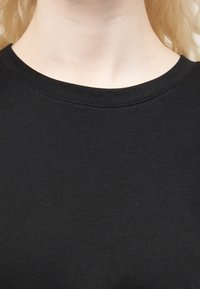 Κοντινή λήψη ενός ατόμου που φοράει απλό μαύρο μπλουζάκι με στρογγυλή λαιμόκοψη, που δείχνει τον λαιμό και την πάνω περιοχή του στήθους.