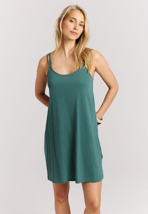 Blonde vrouw met los haar, gekleed in een mouwloze, teal kleurige, knielange jurk met dubbele schouderbanden, staand met haar handen achter haar rug.