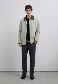 Joven de pie en interiores, vistiendo una chaqueta beige con cuello marrón, camiseta de polo negra, pantalones gris oscuro y zapatos negros, con las manos en los bolsillos.