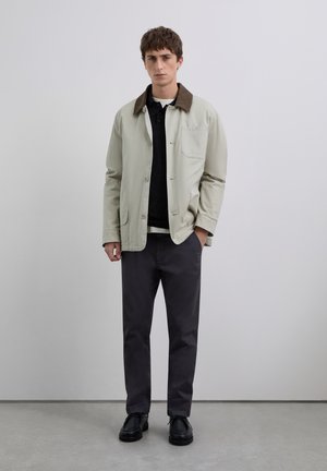 Joven de pie en interiores, vistiendo una chaqueta beige con cuello marrón, camiseta de polo negra, pantalones gris oscuro y zapatos negros, con las manos en los bolsillos.