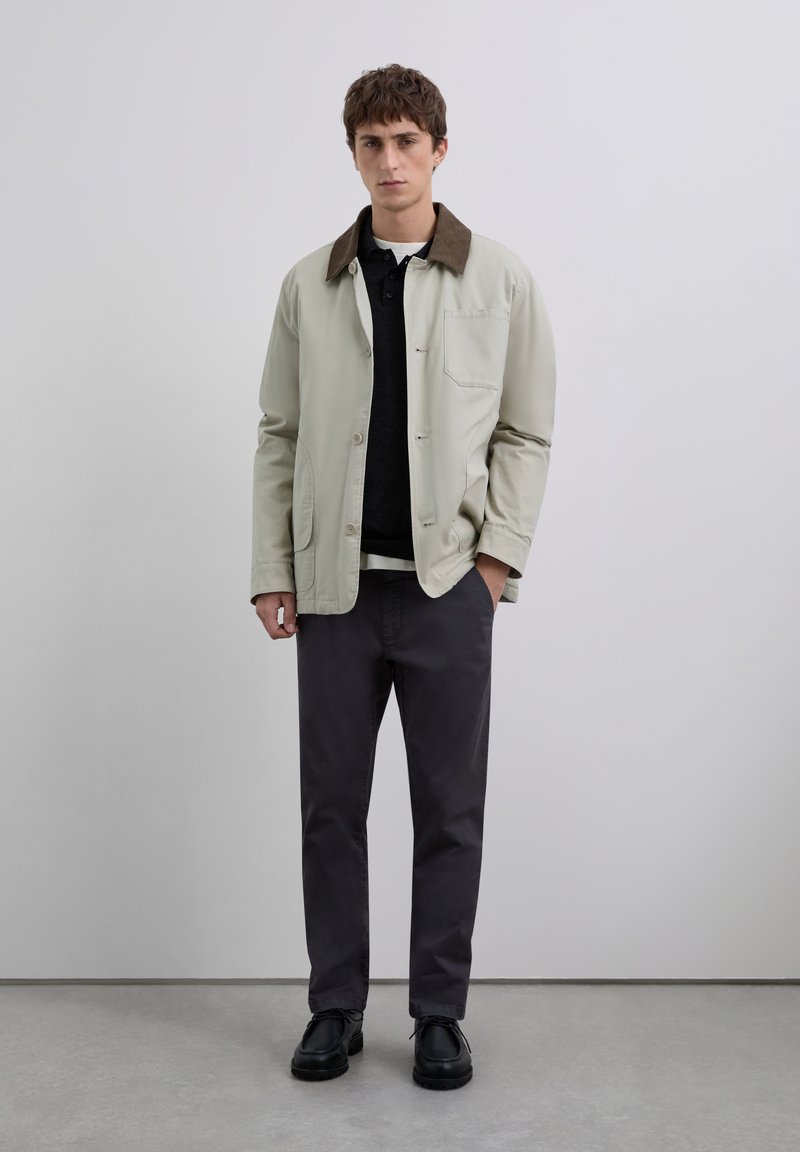 Joven de pie en interiores, vistiendo una chaqueta beige con cuello marrón, camiseta de polo negra, pantalones gris oscuro y zapatos negros, con las manos en los bolsillos.