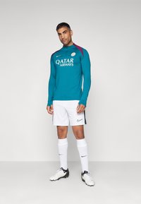 Teal pitkähihaainen urheilu toppi, jossa on korkea kaulus, violetit yksityiskohdat ja valkoinen "QATAR AIRWAYS" -logo, yhdistettynä valkoisiin shortseihin ja mustiin kengillä.