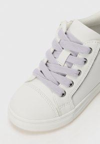 Hvit sneaker med teksturert lær, rund tå og lavendel snørebånd. Har metalliske øyer og en gummisåle med teksturert grep.