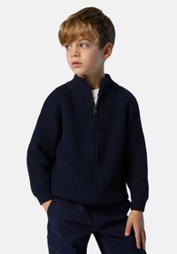 Suéter de punto acanalado azul marino con cremallera y alto cuello, tejido texturizado y mangas largas, combinado con pantalones cargo oscuros.