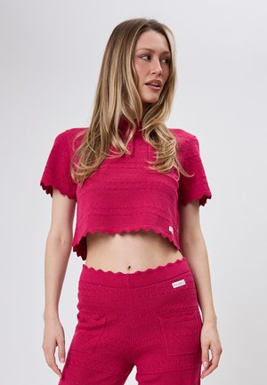 Woman wearing a matching bright pink textured knit crop top and pants set, looking to her right against a plain background.