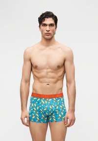 Homme aux cheveux foncés et à la carrure musclée portant un boxer bleu avec des motifs de bananes jaunes et une ceinture rouge.