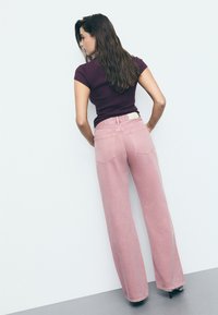 Široke ružičaste traperice viskog struka s pet dzepova, u kombinaciji s pripijenom ljubičastom majicom. Glatka tekstura i minimalna detaljna šavova.