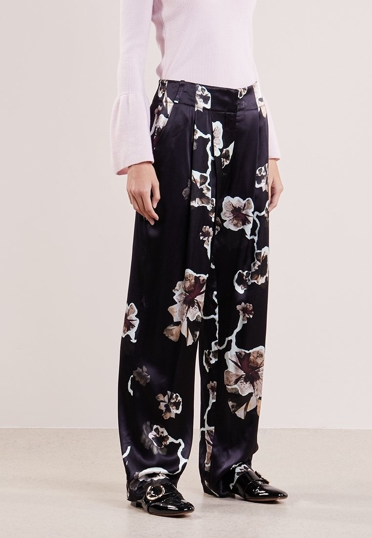 Femme portant un pantalon large en satin noir avec un grand imprimé floral blanc et marron, associé à un haut à manches longues rose clair et des chaussures noires.