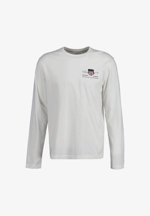 Hosszú ujjú, könnyű anyagból készült fehér ing. Kék és piros nyomtatott logó található a mellrészen, valamint kerek nyakú kialakítás.