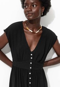 Robe noire texturée à manches courtes, décolleté en V profond et devant boutonné. Accentué par un collier en perles chunk.