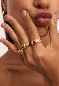 Acercamiento de manos con tres anillos de oro que presentan piedras rojas, verdes y naranjas, presionadas contra labios con esmalte de uñas rosa.
