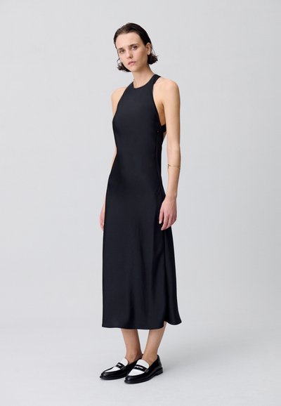 Robe noire sans manches avec une texture lisse, forme A-line et découpes sur les côtés. Associée à des mocassins plats noir et blanc.