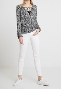Blusa floreale bianca e nera con maniche lunghe, dettaglio del collo a fiocco, abbinata a jeans skinny bianchi e sneakers bianche.