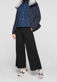 Osoba v námořnické péřové bundě s kožešinovým límcem, modré džíny s knoflíky, širokých černých kalhotách a bílých teniskách s námořnickými a burgundskými akcenty.