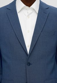 Blazer bleu texturé avec des revers pointus et une fermeture à un bouton, porté sur une chemise blanche à col avec des boutons.