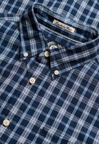 Chemise à carreaux bleu et blanc avec boutons beige, col boutonné et étiquette "Laundered Wash" à l'intérieur du col.