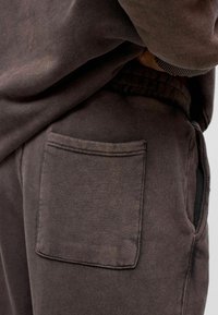 Bruine katoenen sweatpants hebben een enkele achterzak, zichtbare stiksels en een zachte textuur met een lichte kleurvariatie.