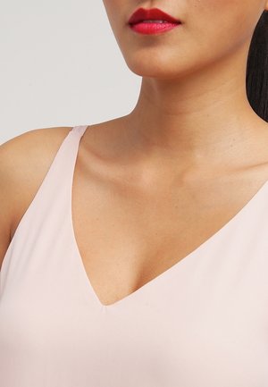 Vrouw met een lichtroze mouwloze top met V-hals, felrode lippenstift, waarbij haar hals, sleutelbeen en een deel van haar schouder zichtbaar zijn.