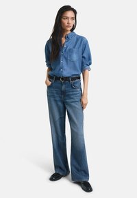 Denim shirt with button-up collar, front pocket, loose fit; paired with high-waisted, wide-leg blue jeans and black shoes.