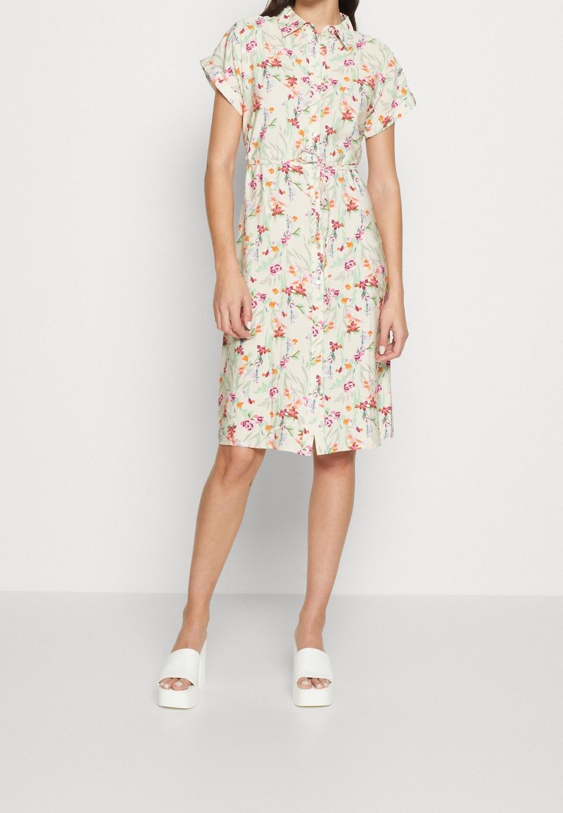 Femme portant une robe à manches courtes, à boutons et ceinture, longueur genou, avec une base blanche et un imprimé floral multicolore, assortie à des sandales à plateforme blanches.