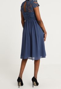 Kvinna klädd i knälång marinblå klänning med spetsöverdel och chiffongkjol, med svarta högklackade skor, står med ryggen mot kameran.