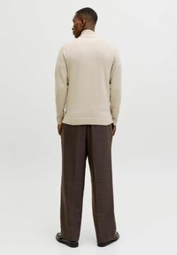 Homme de dos portant un pull à col roulé beige, un pantalon large à carreaux marron, des chaussures noires, et une petite boucle d'oreille sur un fond blanc.