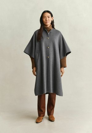 Vrouw draagt een grijze cape met knopen over een bruine coltrui, bruine broek en beige enkellaarsjes, staand tegen een eenvoudige neutrale achtergrond.