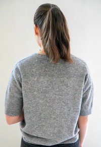 Femme aux longs cheveux bruns en queue de cheval, portant un pull court en tricot gris à manches courtes et un pantalon noir, se tenant contre un fond clair uni.