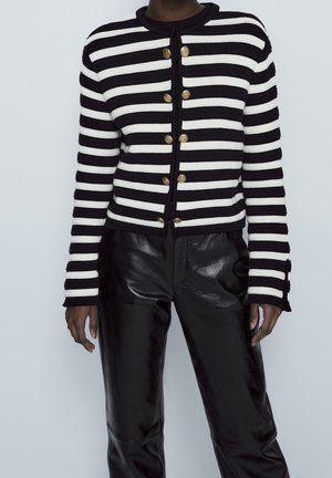 Personne portant un pull à rayures horizontales noir et blanc boutonné avec des boutons dorés et un pantalon en cuir noir brillant.