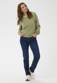 Žena oblečená v olivovo zelenom svetríku, tmavomodrých džínoch a bielych teniskách, stojí so založenými rukami v kapsách na bielom pozadí.