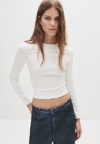 Fiatal nő hosszú barna hajjal, aki egy testhez simuló, fehér, hosszú ujjú crop topot és high-waisted sötétkék farmert visel, egy sima fehér háttér előtt.