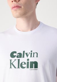 Hvit bomulls t-skjorte med et fremtredende grønt "Calvin Klein" logo trykk. Enkel design med rund hals og glatt stofftekstur.