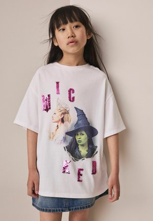 Jong meisje met lang donker haar draagt een wit oversized shirt met twee heksenfiguren en roze paillettenletters die "WICKED" spellen.
