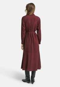 Burgunderfarbenes Langarmkleid mit schmaler Taille und fließendem Rock, gefertigt aus einem weichen Material. Verfügt über Knopfdetails an den Manschetten.