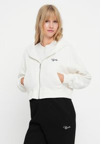 Weißer, verkürzter Hoodie mit Reißverschluss, weichem Material, Fronttaschen und schwarzem gesticktem Logo. Kombiniert mit schwarzen, gerippten Hosen.