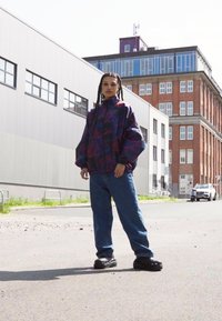 Windbreaker jacket with a colorful abstract pattern, oversized fit, and elastic cuffs. Worn with loose blue jeans and black footwear.