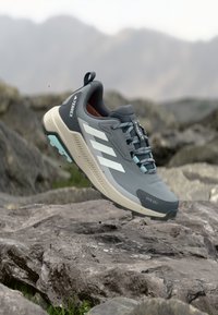 Grauer Trailrunning-Schuh mit Obermaterial aus Mesh und synthetischem Material, mit weißen Streifen, beigefarbener Zwischensohle und türkisfarbenen Akzenten. Profilierte Außensohle für besseren Grip.