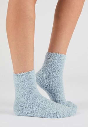 Pieds portant des chaussettes courtes duveteuses bleu clair sur un fond neutre uni.