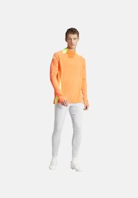Langarm sportshirt in feloranje met gele accenten, voorzien van een gestructureerd oppervlak en hoge kraag. Gecombineerd met witte broek en voetbalschoenen.