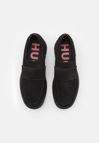 Mocassins en daim noir avec une semelle plate et une large sangle à l'avant. L'intérieur présente des semelles de marque rouges avec une écriture en gras.