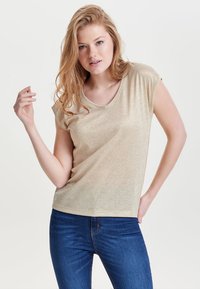 Gul, kortærmet V-hals t-shirt med en blød, skinnende tekstur. Parret med blå denim jeans. Modellen poser med en hånd hævet.