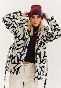 Prošívaná bunda s černobílým květinovým vzorem, prošívaný design, boční zavazování, zapínání na zip a vysoký límec. Nošena přes černý top.