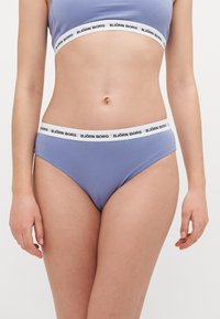 Világoskék bikini alsó, puha textúrával és fehér gumis derékkal, amelyen fekete logó található. Egyszerű dizájn, közepes fedettséggel.