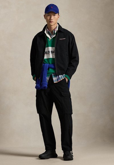 Sort lynlåsjakke, grønstribet sweater, ternet skjorte, blå cardigan bundet om livet, sorte cargobukser, blå kasket, sorte sko.